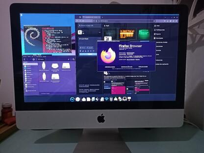 Foto (tomada con un celular) de una iMac de 2010 operando con Elive Linux, escritorio Enlightenment 16 con el tema black-e (marcos de ventana muy delgados, con barra de título recortada como si fuera una pestaña que ocupa la mitad del ancho de la ventana, en color negro con texto blanco). Las aplicaciones usan el tema GTK retrowave-glow (oscuro en tonalidades de púrpura). Hay una ventana de terminal zsh con fondo negro semitransparente mostrando la salida de fastfetch. Debajo se ve una ventana del gestor de archivos Thunar, mostrando las unidades de almacenamiento del equipo, entre las que se ven las típicas de iMac. A la derecha una gran ventana de Firefox mostrando mi perfil en Mastodon y encima la ventana Acerca de Firefox, indicando la versión 136.0.3 instalada. Al pie de la pantalla se ve Cairo-Dock con varios iconos de menú y lanzadores.